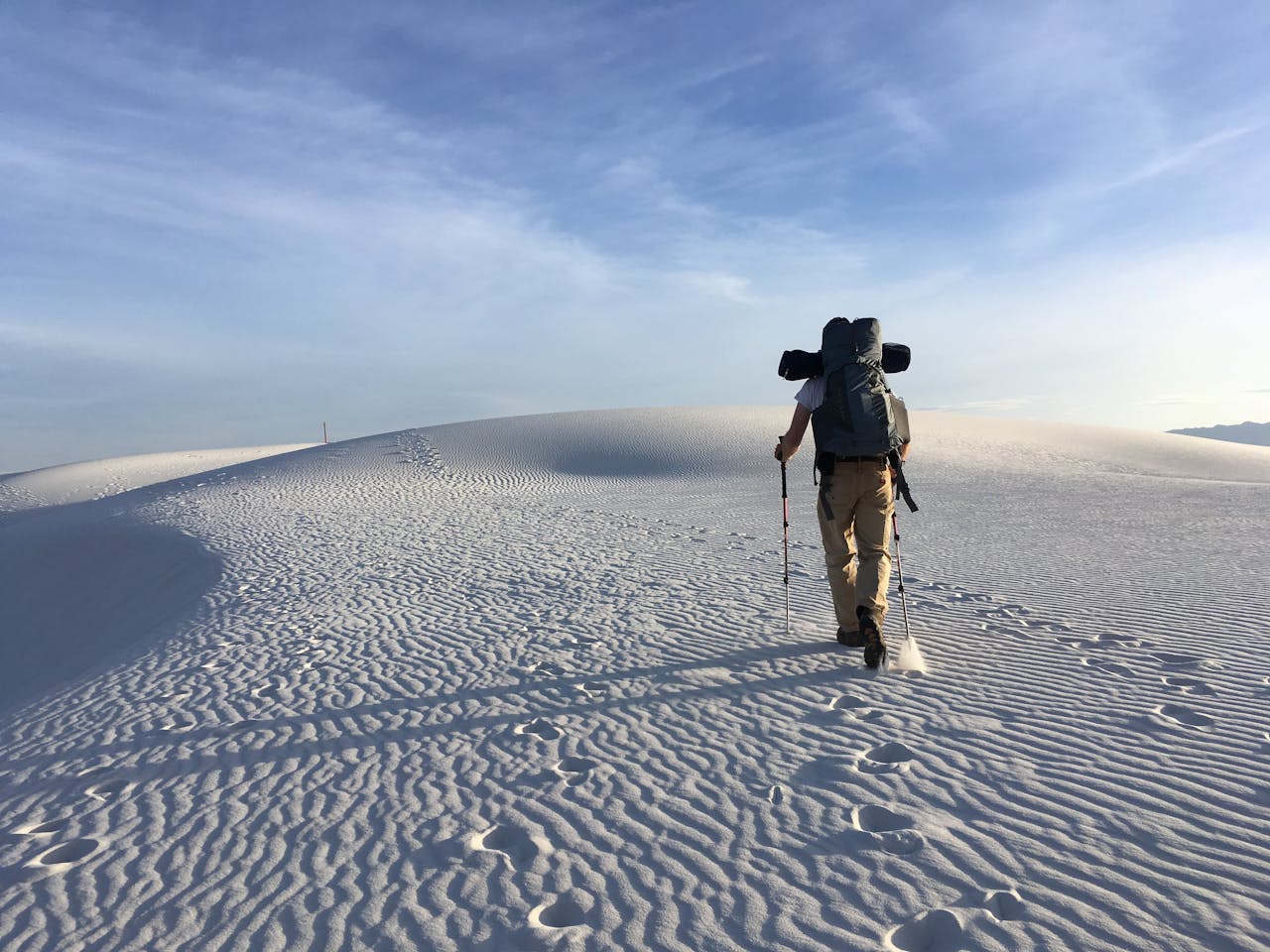 A lone hiker traverses the white sand dunes of White Sands National Park under a clear sky.
