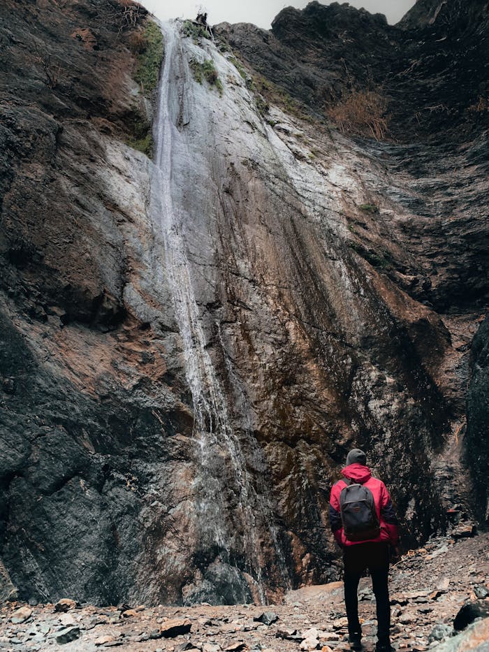 A lone hiker in a red jacket stands before a towering waterfall in rocky terrain.
